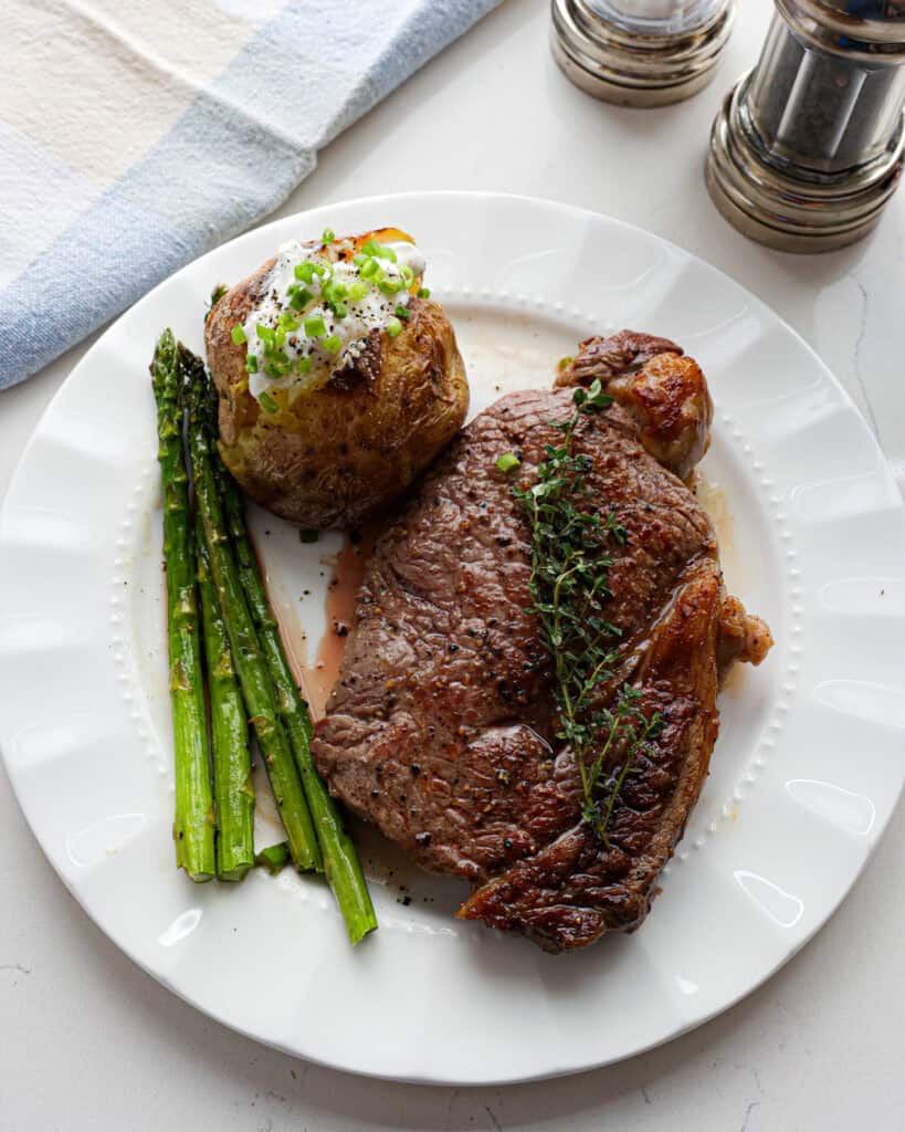 A cast irons skillet steak on a white serving plate with asparagus and a baked potato with souir cream and green onions.