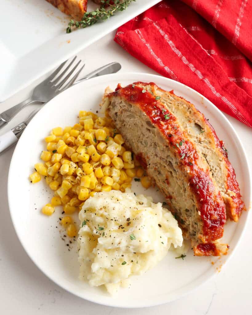 Two slices of glazed chicken meatloaf on a single serving plate with mashed potatoes and corn.