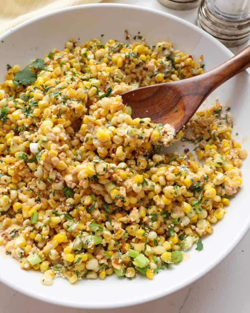 A white serving bowl and a wooden spoon full of Mexican street corn salad.