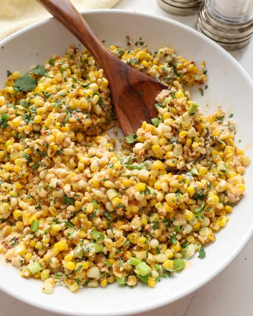 A bowl and a wooden spoon full of elote salad with a salt and pepper shaker in the background.