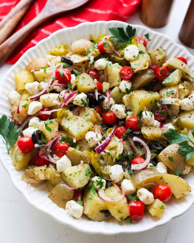 A close up view of Italian potato salad in a white serving bowl with serving tongs in the background.