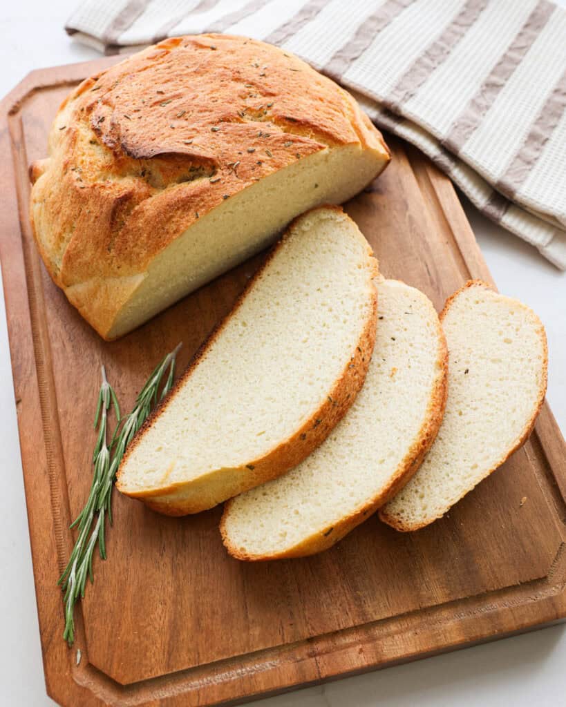 A sliced loaf of rosemary potato bread on a cutting board with fresh rosemary.