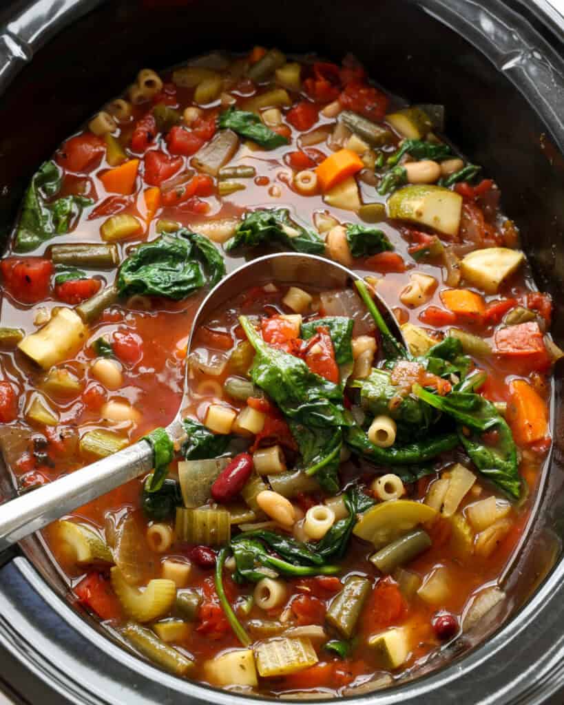 A close up view of a crock pot and ladle full of minestrone soup with carrots, zucchini, tomatoes, pasta, green beans, and beans.