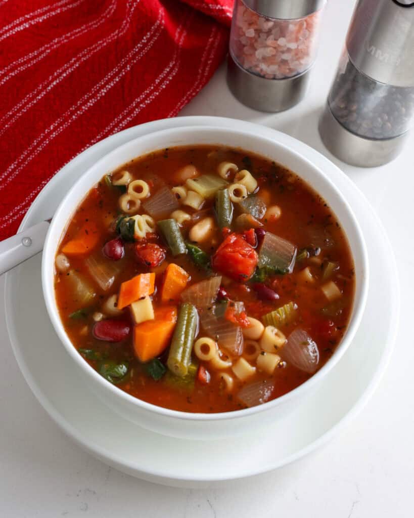 A bowl full of slow cooker minestrone soup with a red kitchen towel and a salt and pepper shaker in the background.